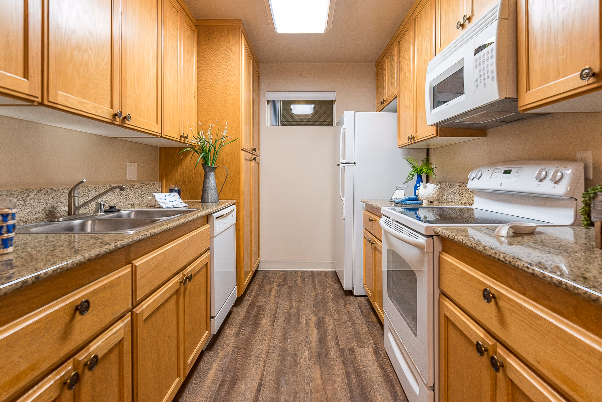 A narrow kitchen with wooden cabinets on both sides, granite countertops, a white refrigerator, white microwave, white electric stove, and a double sink. The floor is wood-patterned, and there are small decorative plants on the counters.
