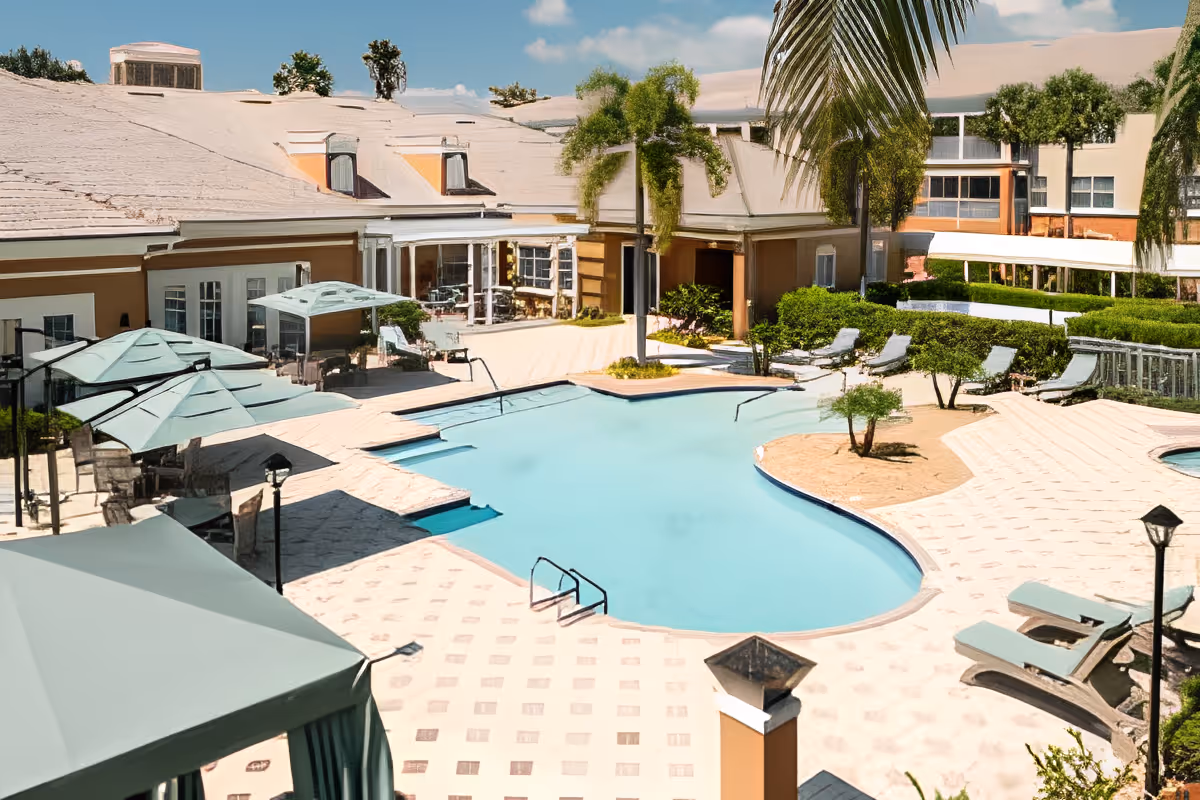 Outdoor swimming pool area at Aston Gardens At Pelican Marsh with lounge chairs, umbrellas, palm trees, and a building in the background under a blue sky.