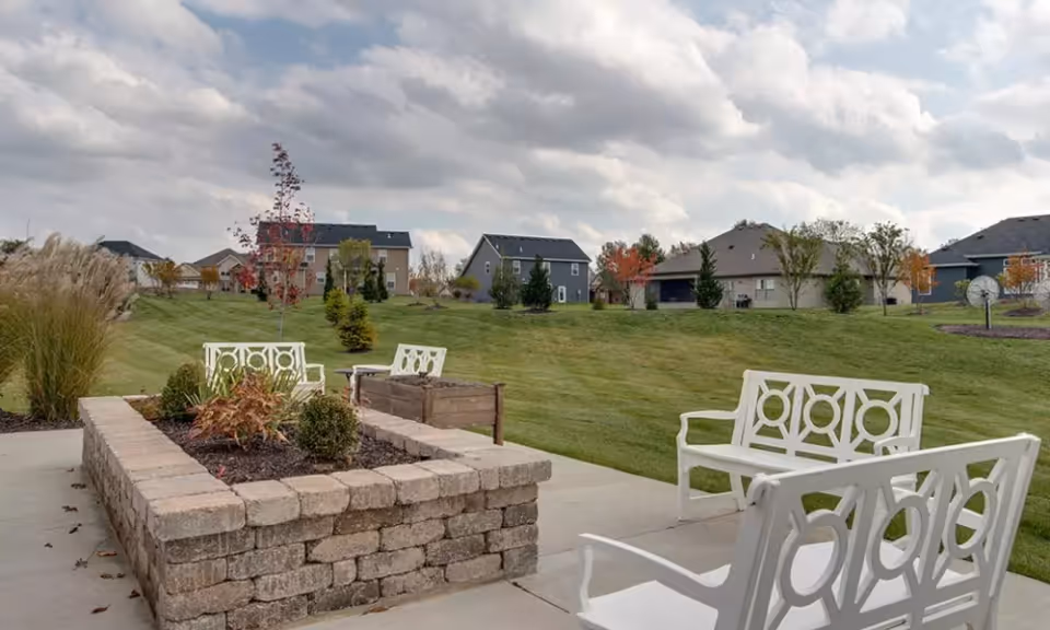 Outdoor patio with white metal benches around a raised brick planter overlooking a grassy lawn and nearby houses under a cloudy sky.