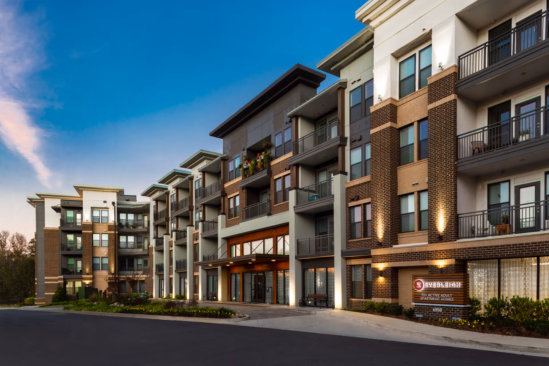 Exterior view of a modern multi-story apartment building with balconies, well-lit entrance, and landscaped surroundings under a clear blue sky at dusk.