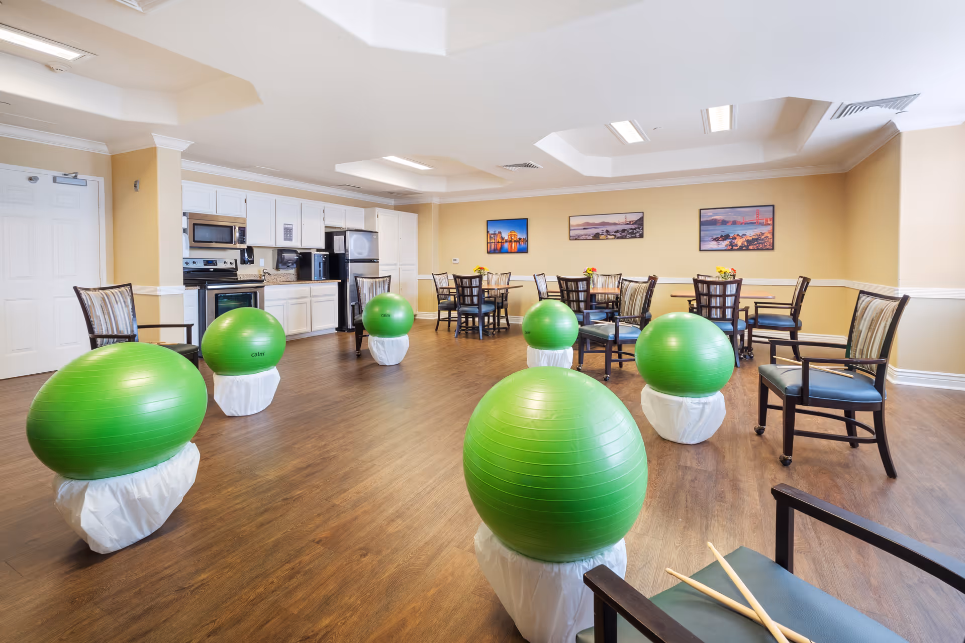 A spacious room with wooden flooring featuring several green exercise balls placed on white stands, surrounded by chairs. In the background, there is a kitchen area with white cabinets, a microwave, stove, and refrigerator. Several tables and chairs are arranged near the walls, and three framed pictures hang on the beige walls.