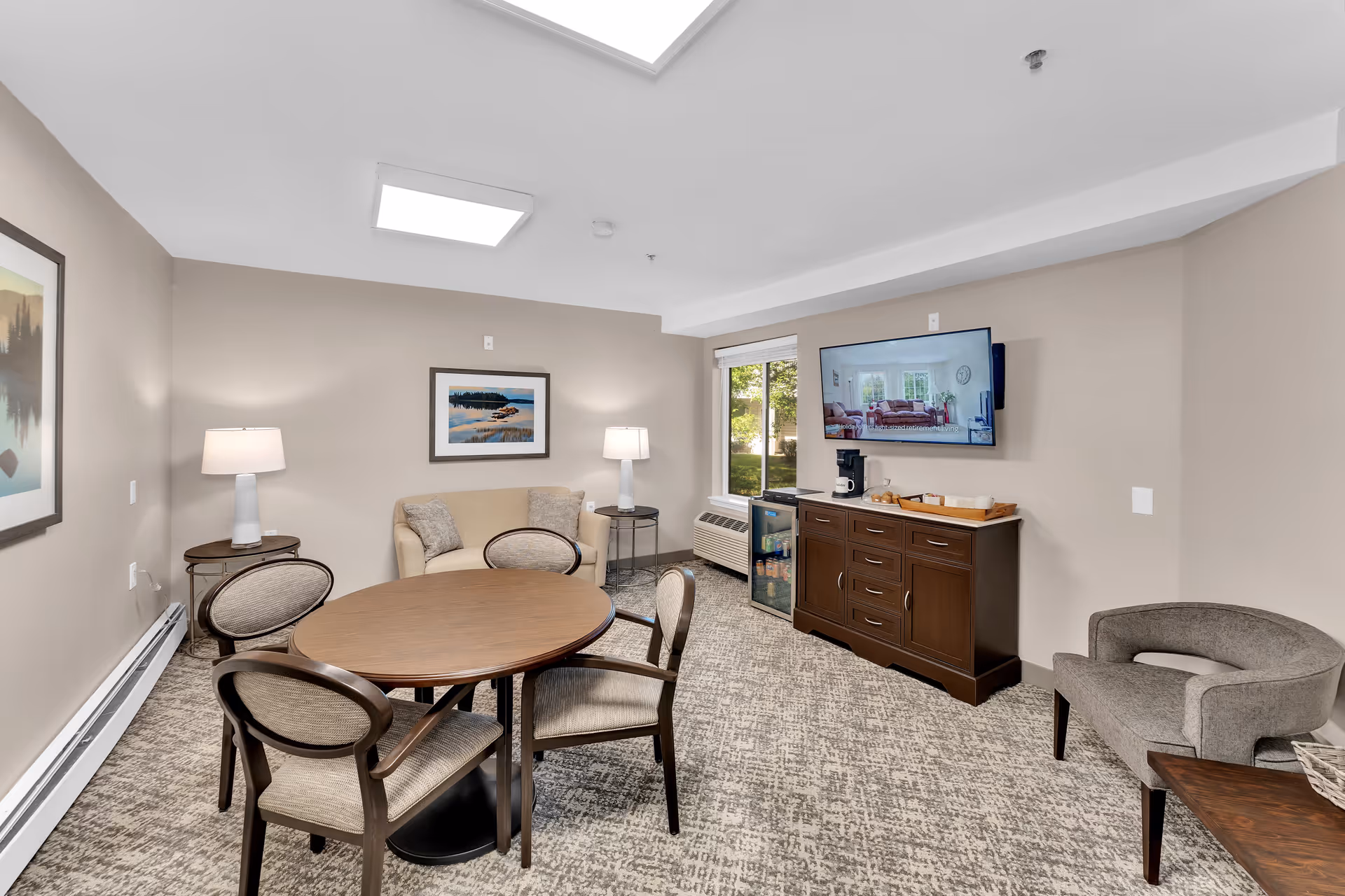 Common room with a round wooden table and chairs, a small seating area with lamps, and a wall-mounted TV above a cabinet.