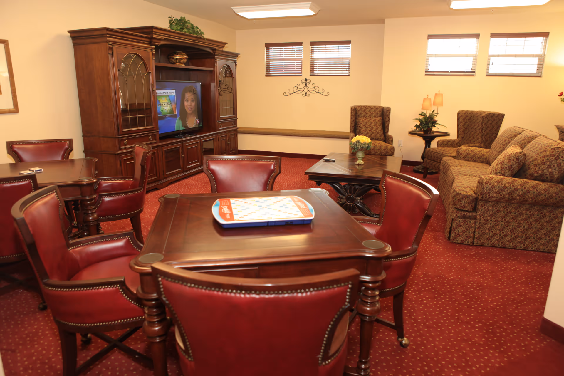 A cozy living room area with a red carpet and cream-colored walls. The room features a wooden entertainment center with a TV showing a woman, a set of four red leather chairs around a wooden table with a board game on it, a patterned sofa, two matching armchairs, a wooden coffee table with a flower vase, and a small side table with a lamp and plant. There are three small windows with blinds near the ceiling.