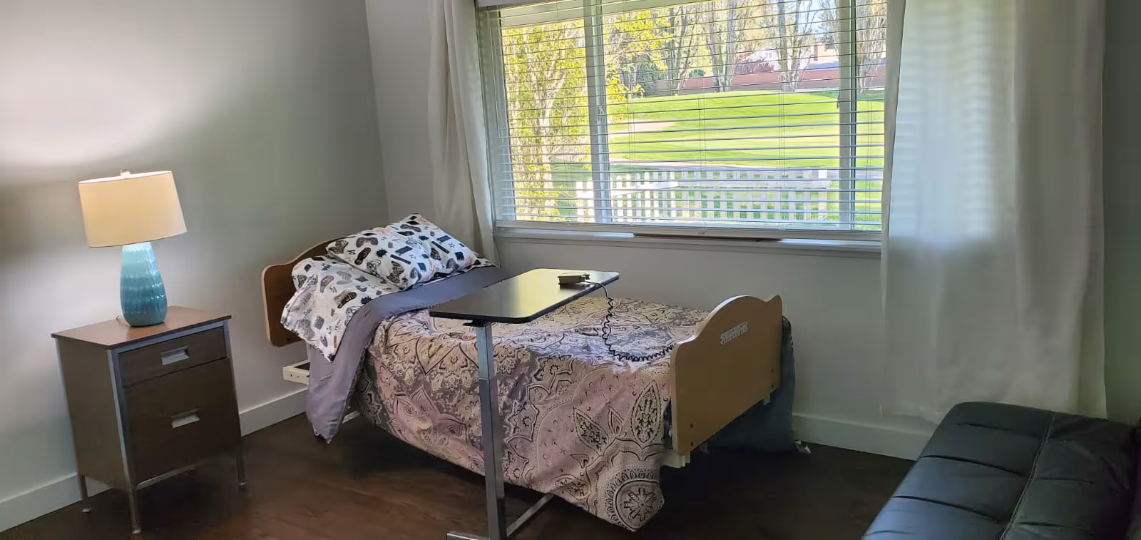 A small bedroom with a single hospital-style bed covered with a patterned bedspread and pillows. Next to the bed is a wooden nightstand with a blue lamp. A window with white blinds shows a green outdoor area with trees and a white fence. There is a black cushioned bench or sofa on the right side of the room.