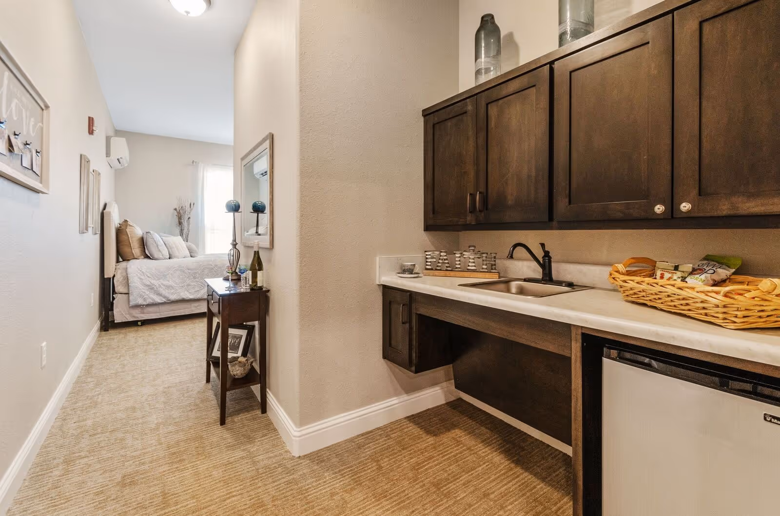 Interior view of a small apartment-style room with a kitchenette featuring dark cabinets and a sink and a bed visible down the hallway.