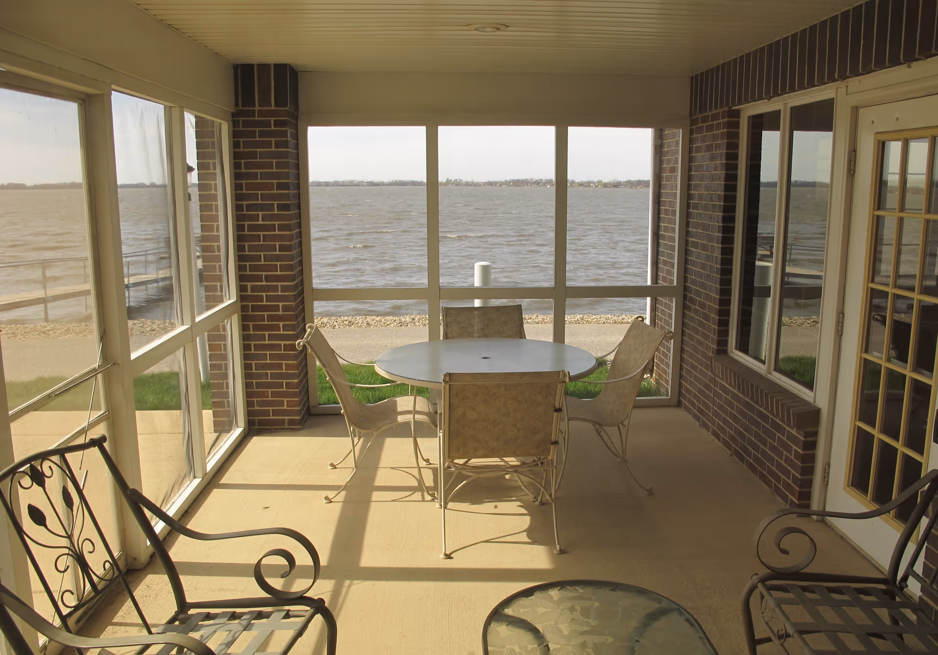 A screened-in porch area with a round table and four chairs in the center, two metal chairs with decorative backs on the sides, and a view of a large body of water beyond the porch. The porch has brick walls and large windows allowing natural light to fill the space.