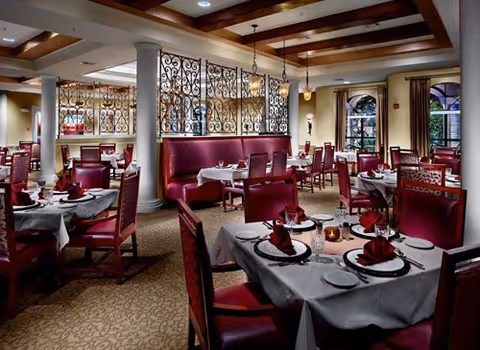 Elegant dining room with tables set for a meal, featuring white tablecloths, folded red napkins, plates, and glassware. The room has red upholstered chairs and booths, decorative wrought iron dividers, carpeted floor, and warm lighting from ceiling fixtures and windows.