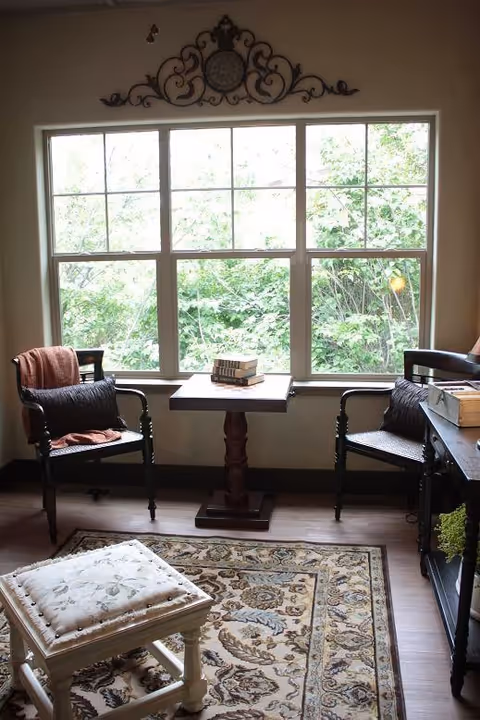 A cozy sitting area with two wooden chairs featuring cushions and a small wooden table between them, placed in front of a large window with a view of green foliage outside. A decorative wall piece is mounted above the window. There is a patterned rug on the wooden floor and a cushioned stool in the foreground.