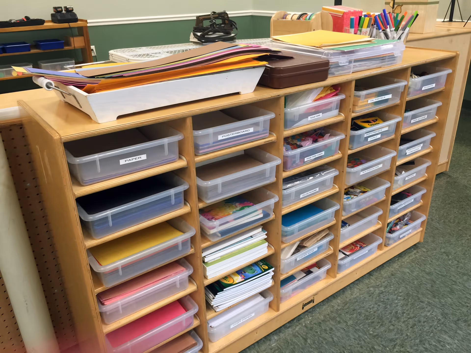 A wooden storage unit with multiple labeled plastic bins containing various art supplies such as paper, poster board, tissue paper, oil pastels, chalk, watercolor, and collage magazines. On top of the unit are trays holding colorful sheets of paper and containers with markers and other craft materials. The room has green walls and a green floor.