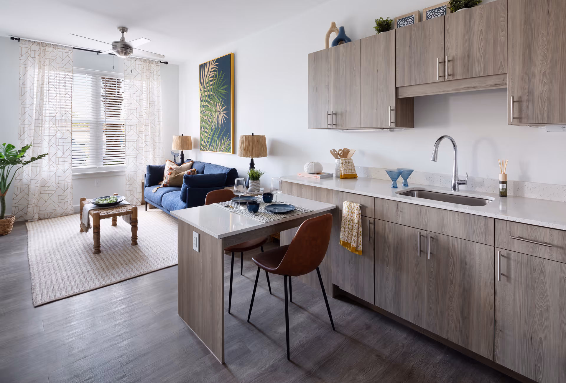 A modern open-concept living space featuring a kitchen area with light wood cabinets, a sink, and a small dining counter with two brown chairs. Adjacent to the kitchen is a cozy living room with a blue sofa, two table lamps, a wooden coffee table on a beige rug, a large window with sheer curtains, and a framed botanical artwork on the wall.