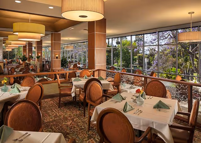 Well-lit dining room with multiple set tables and chairs beside large windows overlooking a garden.