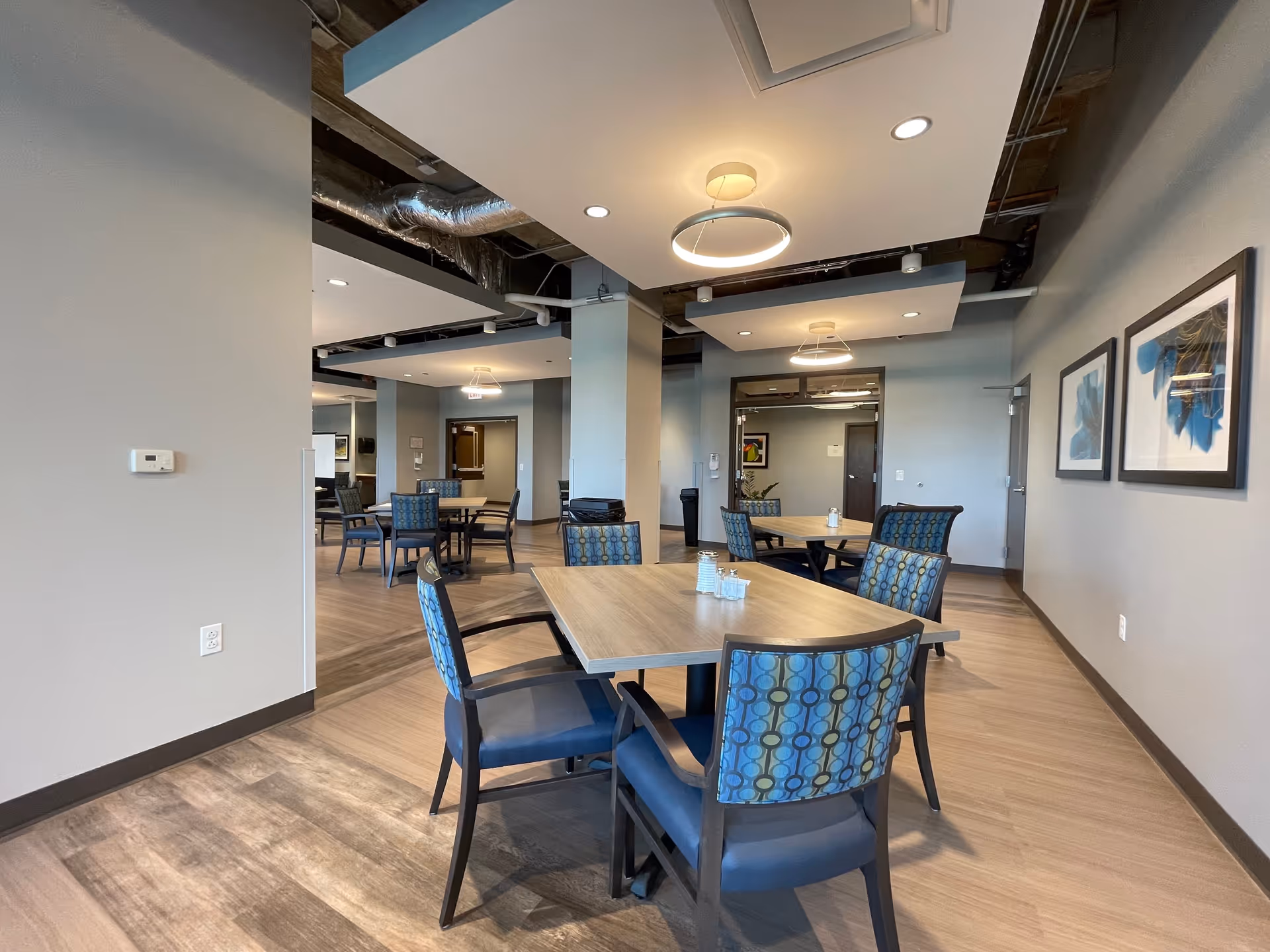Open modern dining area with several wooden tables and patterned chairs in a supportive living facility interior.