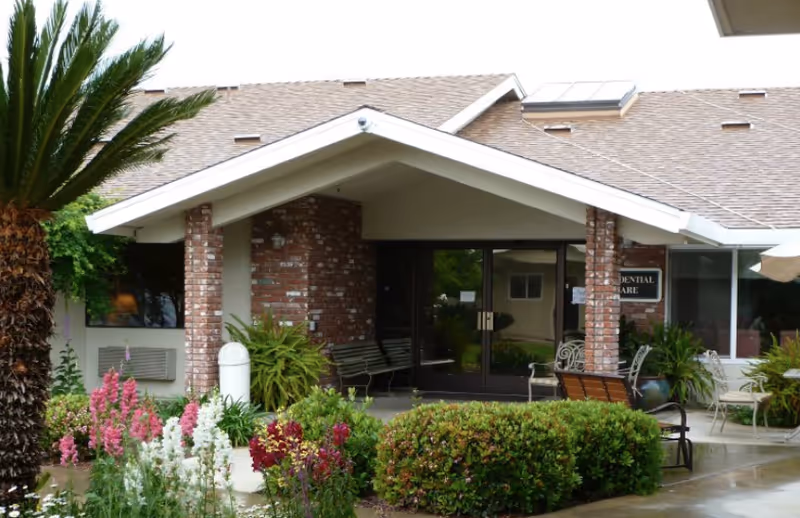 Entrance to a residential care facility with a covered porch supported by brick columns, surrounded by green shrubs and colorful flowers. There are benches and chairs near the entrance, and a sign on the wall reads 'Residential Care'.