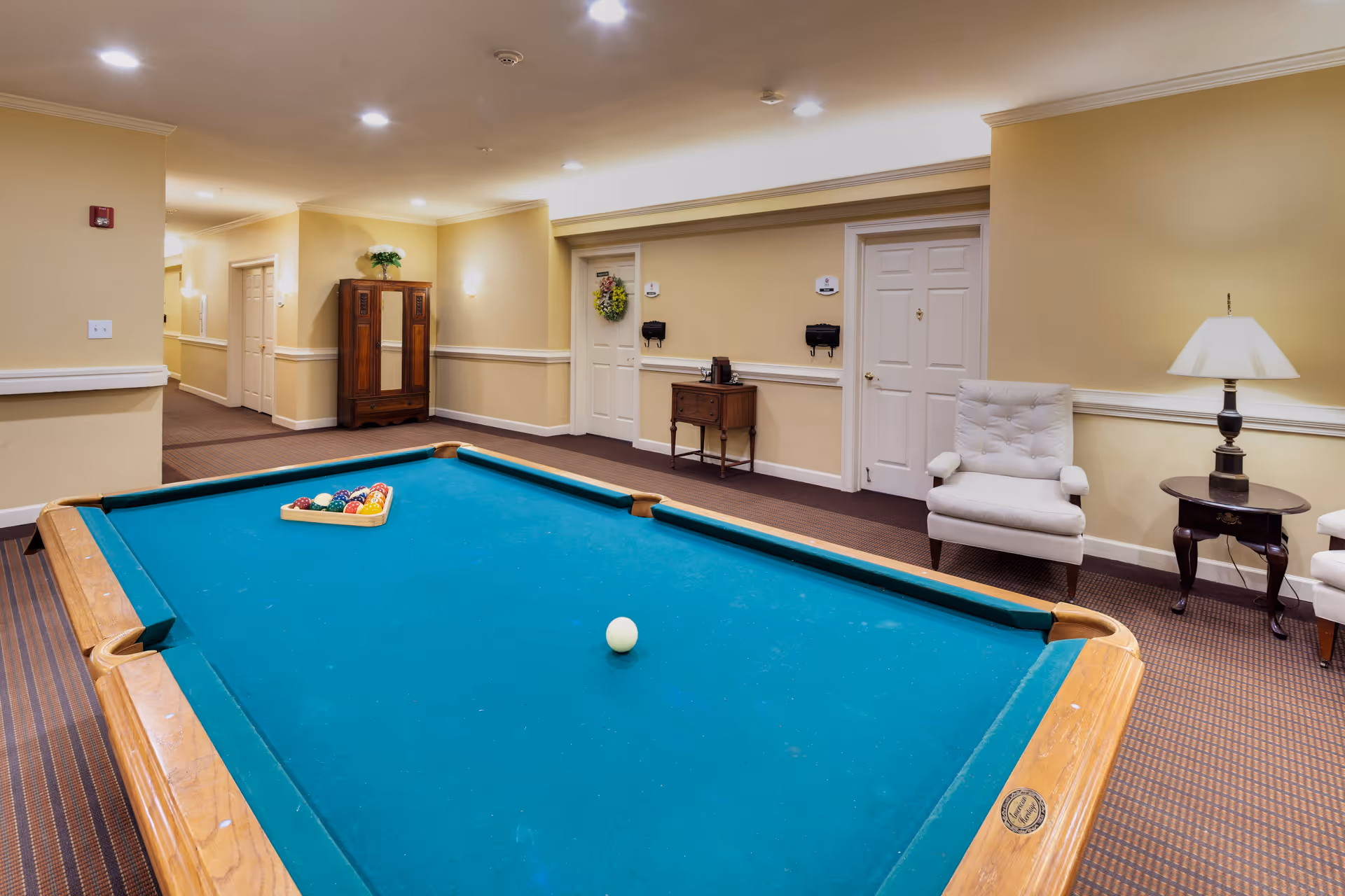 A recreational room with a pool table in the center, set up with billiard balls and a cue ball. The room has beige walls, carpeted floors, two white cushioned armchairs, a small wooden table with a lamp, and a wooden cabinet against the wall. There are two closed white doors and a hallway leading to other rooms.