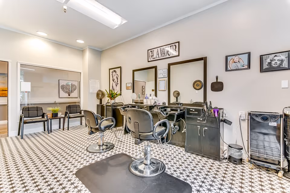 Interior view of a senior living facility's hair salon area with two black salon chairs in front of mirrors and hair washing sinks. The room has patterned tile flooring, framed pictures on the walls including a 'GLAMOUR' sign, and a waiting area with two black chairs and a small table with a plant.