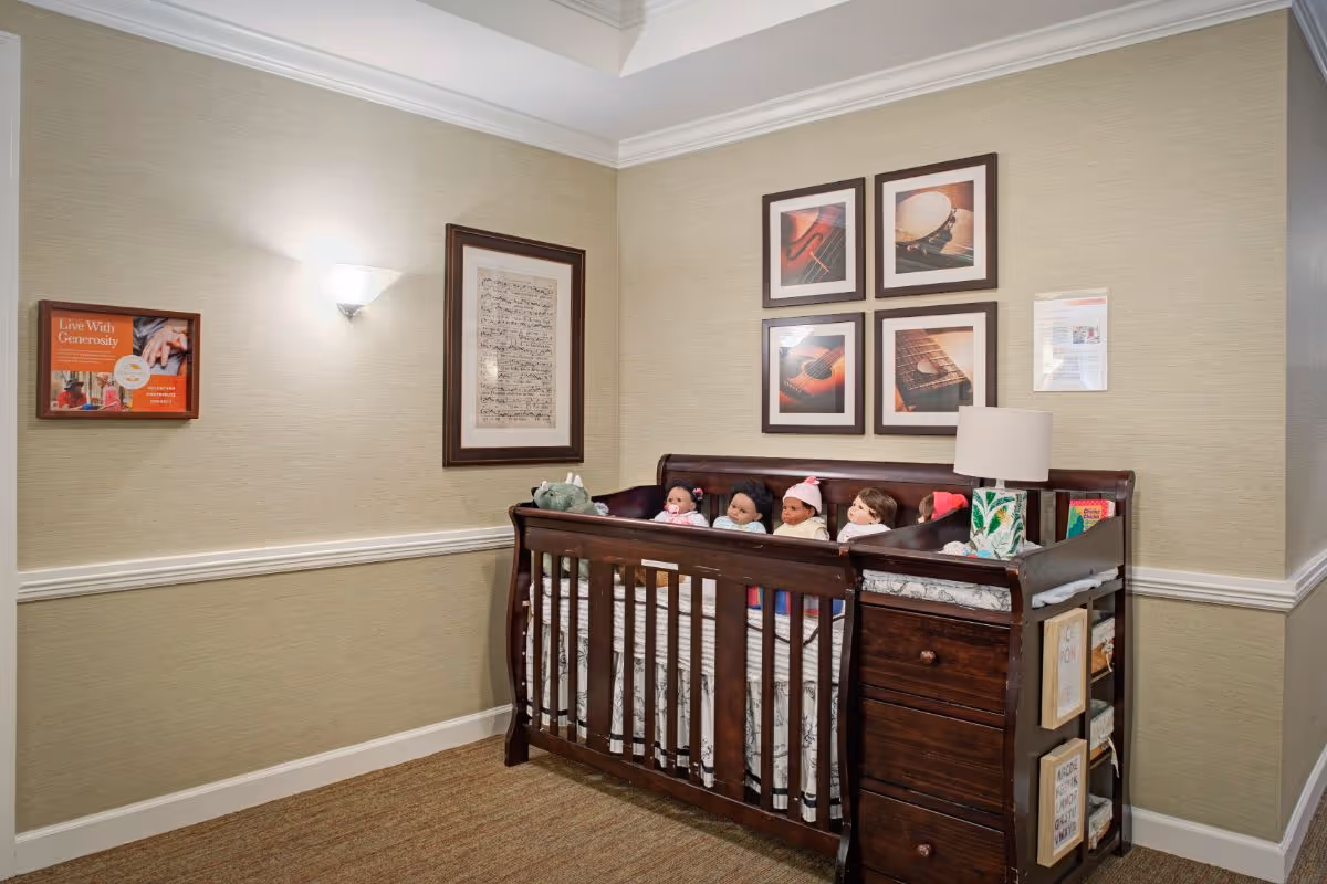 A corner of a room with beige walls and carpeted floor featuring a dark wooden crib filled with several baby dolls. Above the crib are four framed pictures of musical instruments. On the left wall, there is a framed poster titled 'Live With Generosity'. The crib has a built-in changing table with a lamp and some books on it.