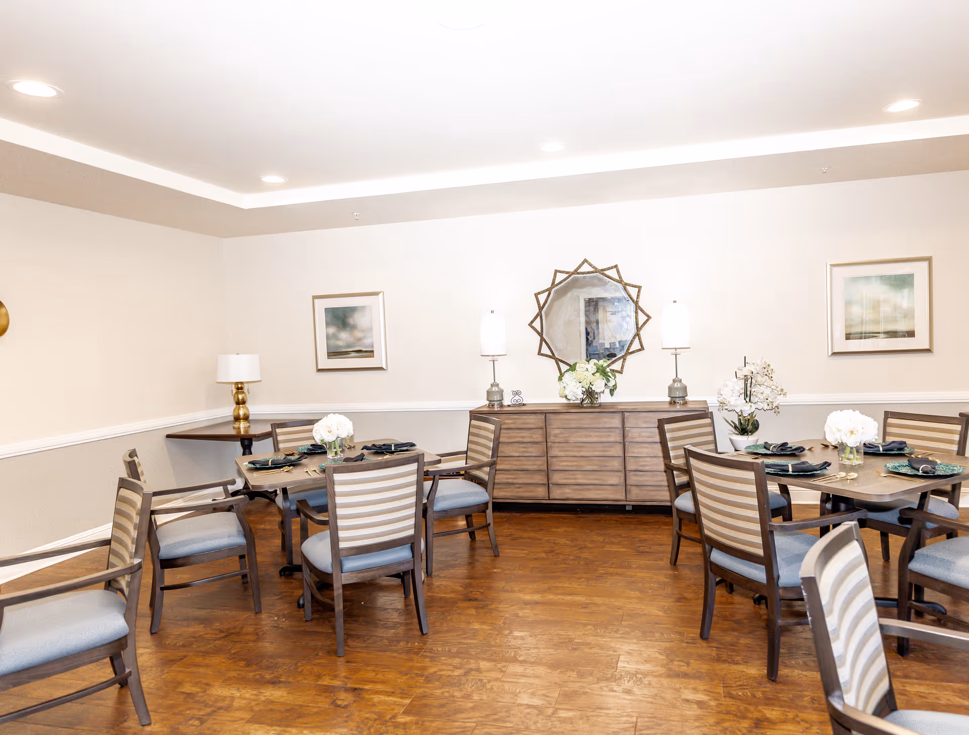 A dining room with wooden floors and several tables set with plates, napkins, and flower centerpieces. The room has beige walls with white trim, two framed pictures, a wooden sideboard with two lamps and a decorative mirror above it.