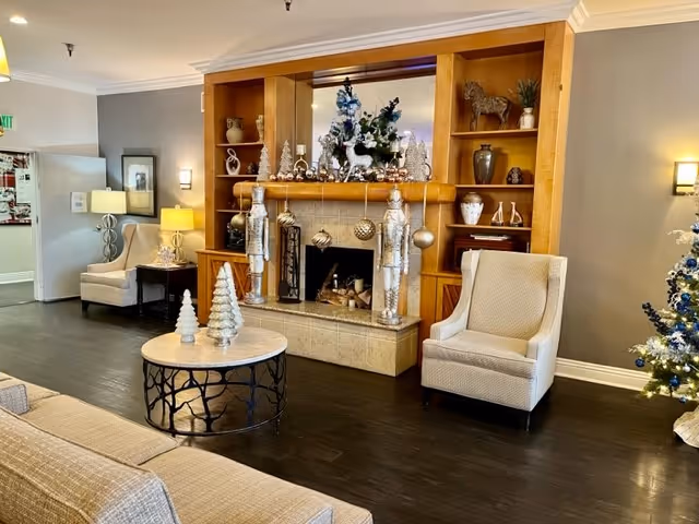 Cozy assisted living lounge with a decorated fireplace, built-in shelves, armchairs, sofa, and a round coffee table with holiday decor.