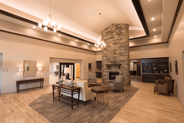Spacious senior living lounge with high ceilings, a central stone fireplace, multiple seating areas and contemporary lighting.