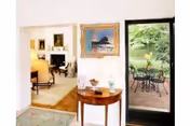 Interior view of a living space showing a small wooden table with decorative items, a framed painting on the wall, a doorway leading to a room with a fireplace and furniture, and a glass door opening to an outdoor patio with a table and chairs surrounded by greenery.