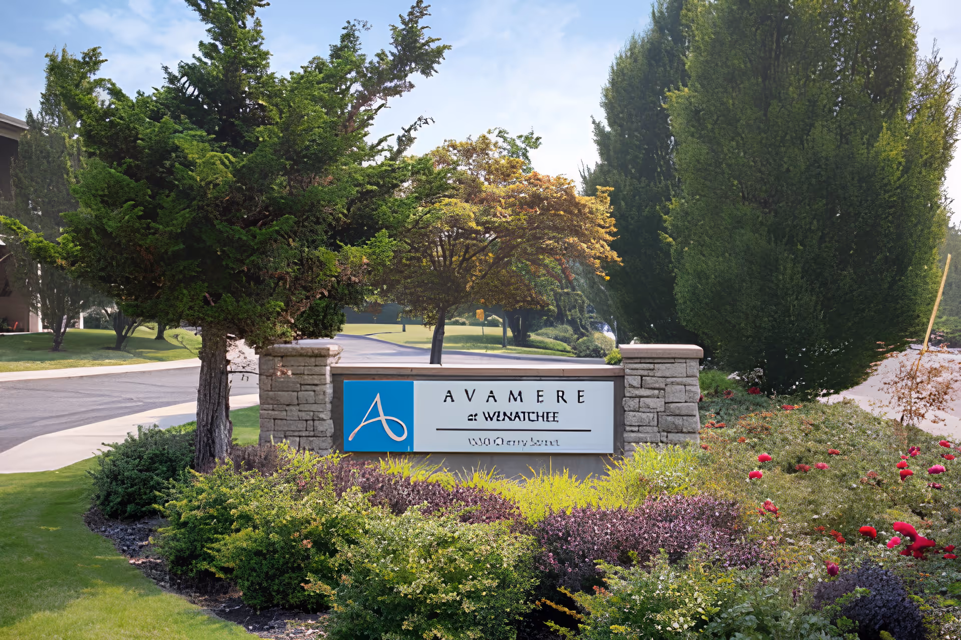 Stone sign at the entrance of Avamere at Wenatchee senior living facility surrounded by green bushes, colorful flowers, and tall trees with a paved road in the background under a clear sky.