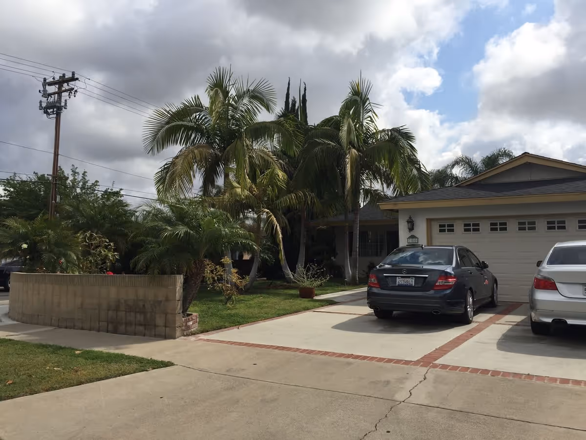 Driveway of a residential home with two parked cars in front of a garage. The house is surrounded by palm trees and other greenery under a partly cloudy sky.