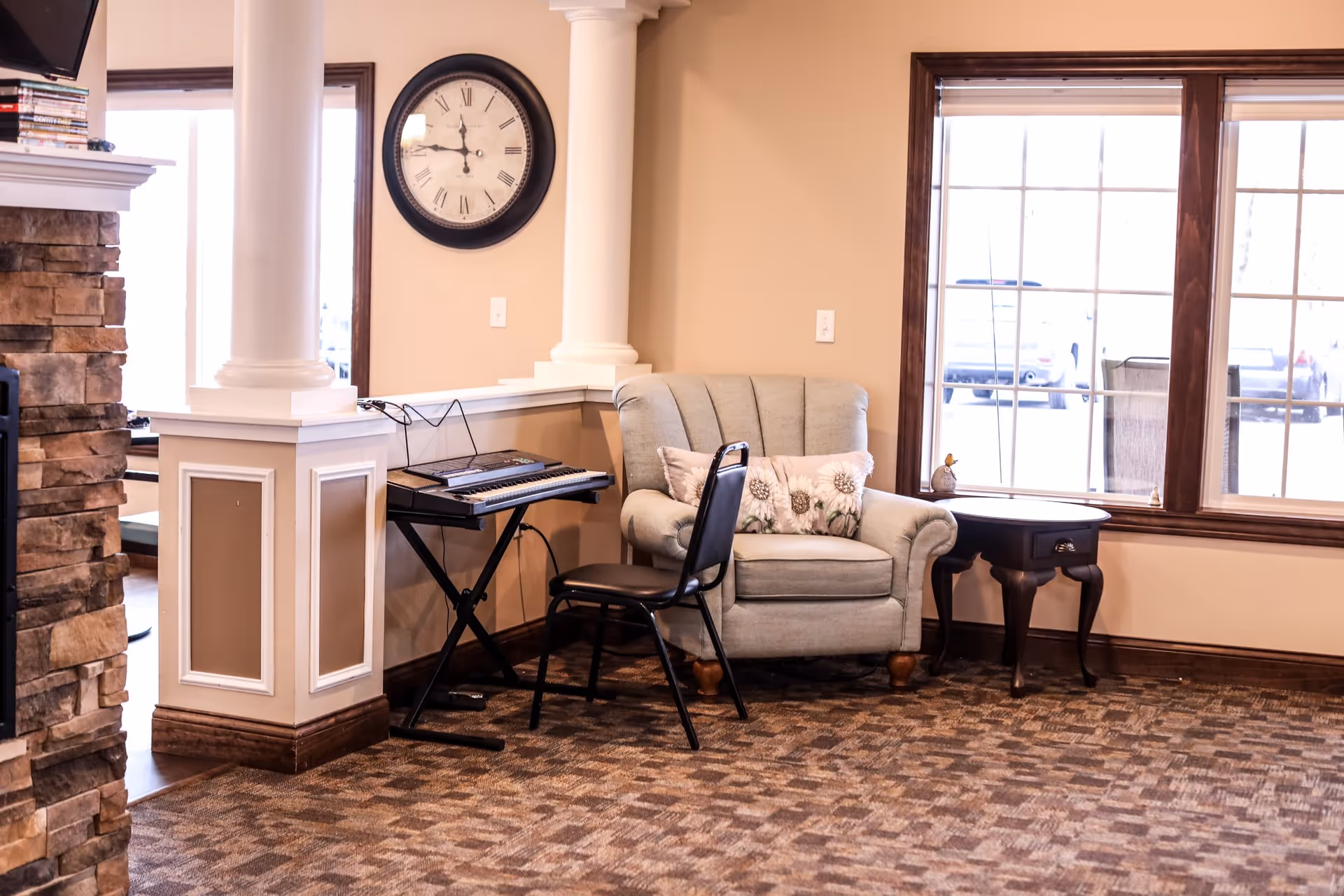 A cozy corner of a living room with a light gray armchair adorned with a floral pillow, a small dark wooden side table with decorative items, a black chair, and an electronic keyboard on a stand. The room features beige walls, a large window with wooden trim, a wall clock with Roman numerals, and a stone fireplace partially visible on the left.