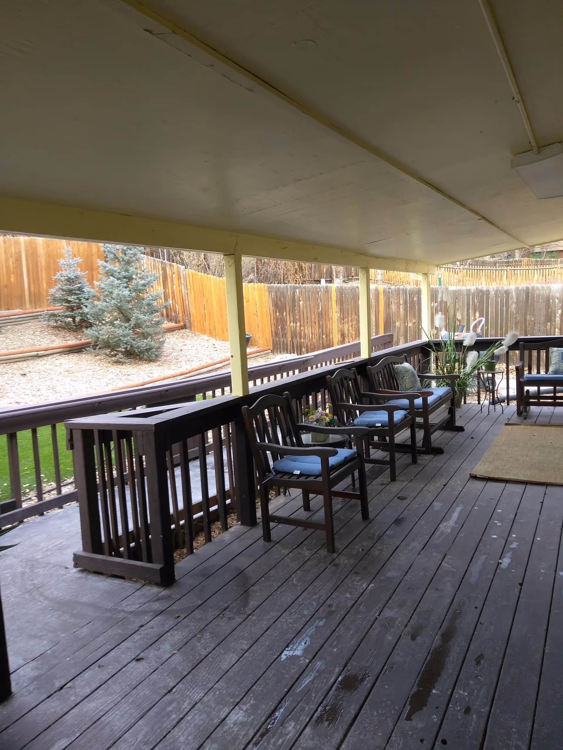 Covered outdoor wooden deck with several wooden chairs with cushions arranged along the railing. The deck overlooks a fenced yard with small evergreen trees and a sloped area covered with mulch.