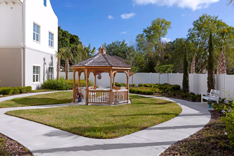 Sunny courtyard featuring a wooden gazebo on a grassy island surrounded by a curved concrete walkway, benches, landscaping and a building.
