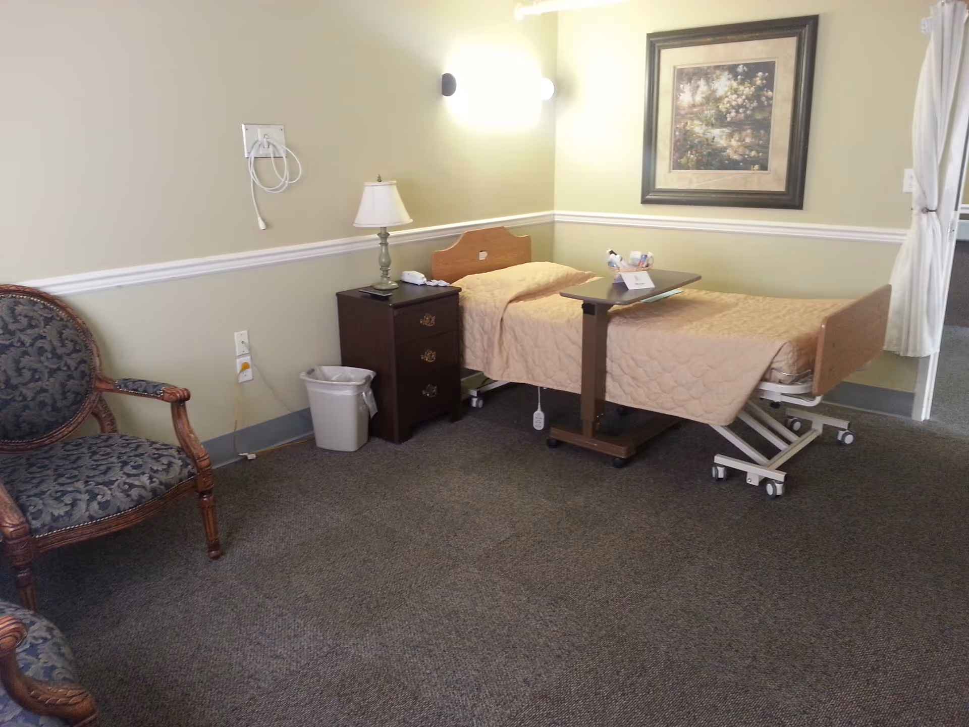 A small, simple bedroom in a senior living facility with a single adjustable bed covered with a beige quilt. Next to the bed is a dark wooden nightstand with a lamp, a phone, and some items on it. There is a trash bin on the floor beside the nightstand. A cushioned armchair with a patterned fabric is positioned to the left. The walls are painted light green with a white chair rail molding, and a framed picture hangs above the bed. A curtain is partially visible on the right side of the image.