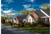 A row of single-story residential cottages with well-maintained lawns and shrubs under a partly cloudy blue sky, with a person walking on the sidewalk beside the houses.