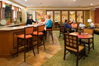 A cozy senior living facility common area with a bar counter on the left where a staff member is serving two seated women. In the background, several seniors are seated at tables and on couches, engaging in conversation. The room is warmly lit with pendant lights, decorated with plants and artwork, and has a mix of tiled and carpeted flooring.