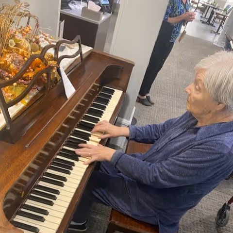 An elderly woman playing a wooden piano in an indoor setting. The piano has autumn-themed decorations on top. Another person is standing in the background near a hallway.