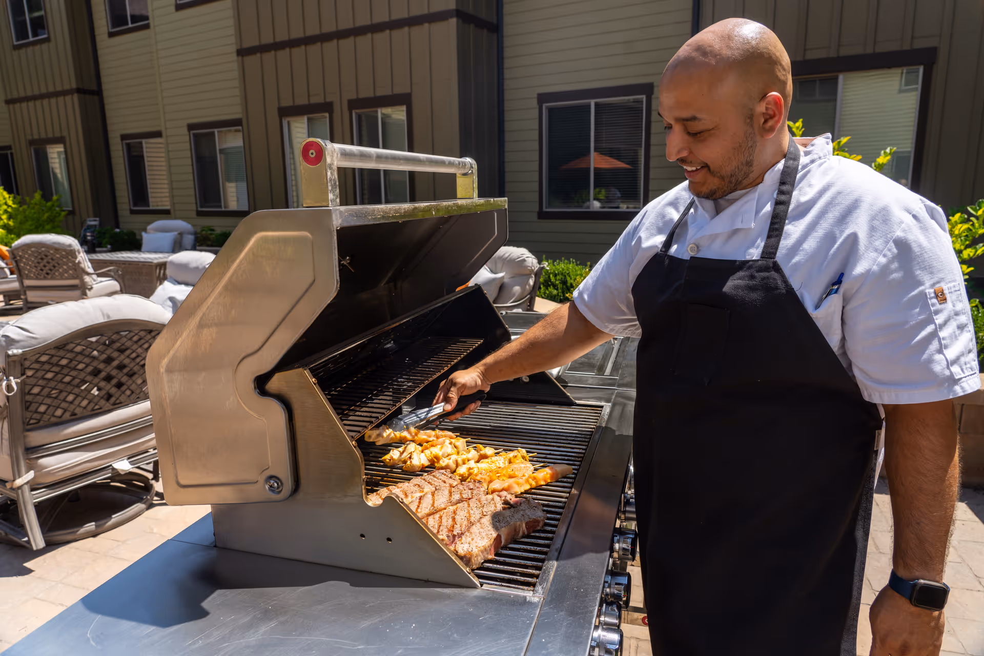 A man wearing a white chef's coat and black apron is grilling food on a large outdoor grill. The grill is open, showing skewers of chicken and a large piece of steak cooking. The setting is an outdoor patio area with cushioned chairs and a building with windows in the background.