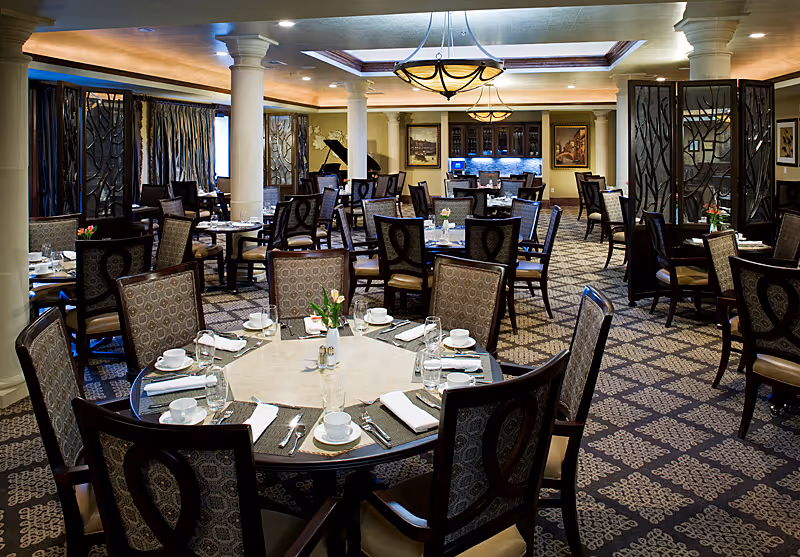 Elegant dining room with multiple round tables set with white cups, plates, napkins, and silverware. The room features patterned carpet, decorative chairs, columns, and warm lighting with chandeliers. There are decorative screens and framed artwork on the walls.
