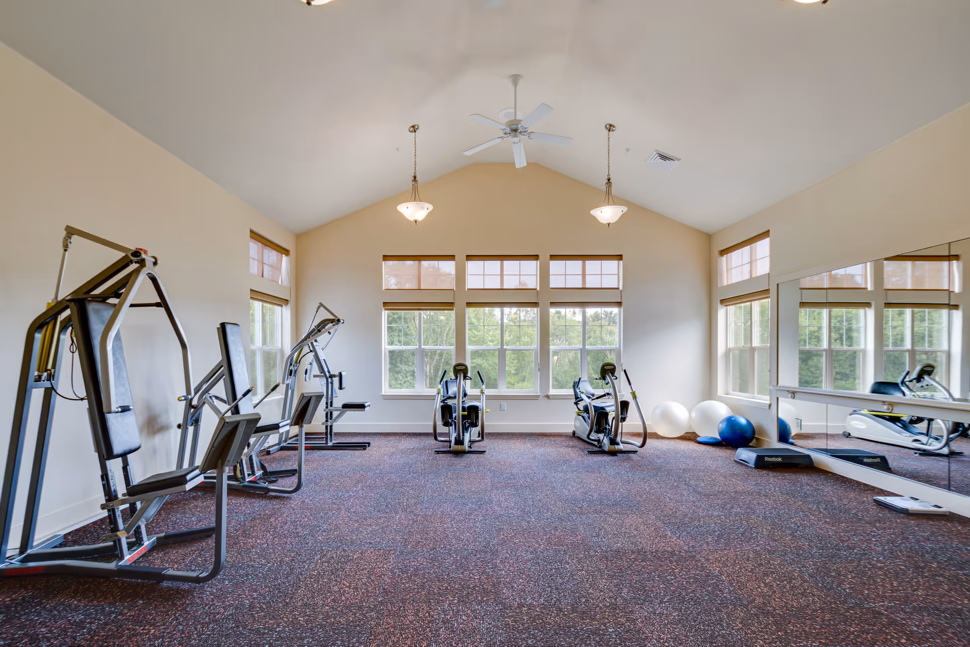 Bright exercise room with large windows, two weight machines on the left, two recumbent exercise bikes in the center, and several exercise balls and a step platform on the right near a large wall mirror. The room has a high vaulted ceiling with a ceiling fan and two hanging light fixtures.