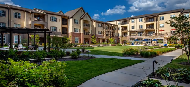 Outdoor courtyard area of a senior living facility with well-maintained green lawns, landscaped bushes, a paved walkway, and a multi-story building with balconies under a blue sky with some clouds.