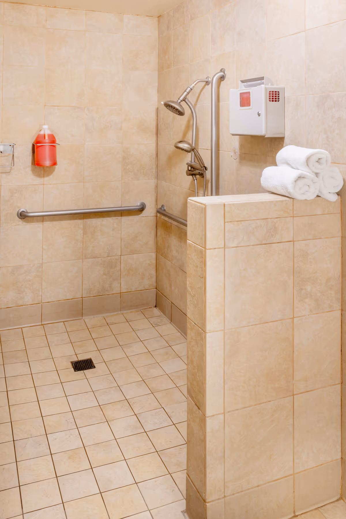 Tiled accessible walk-in shower with grab bars, a handheld showerhead, folded towels on a half wall and an emergency alarm/dispenser.