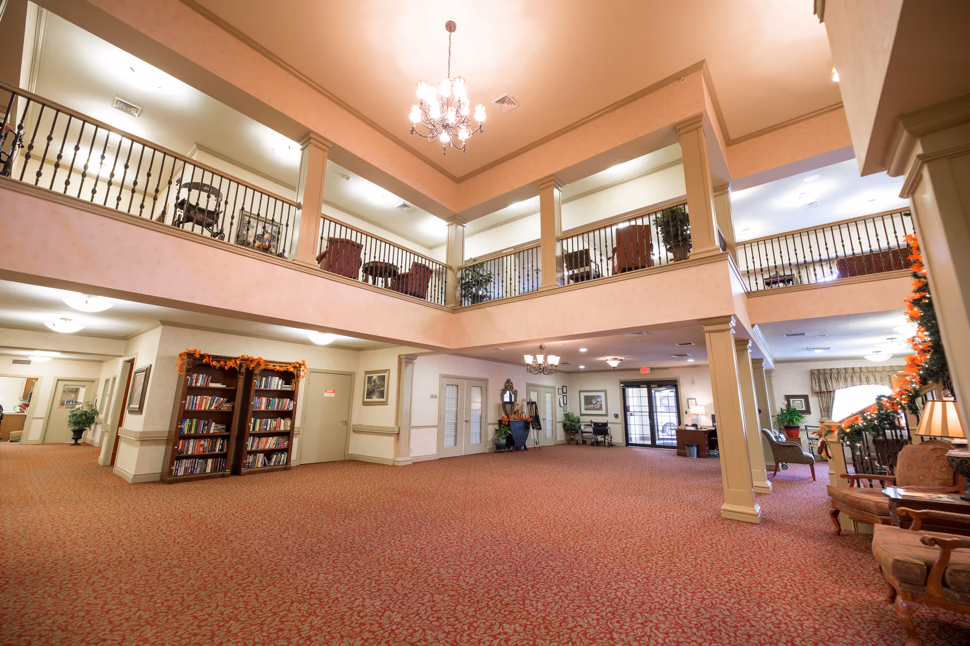 Spacious two-story interior of a senior living facility with red patterned carpet, beige walls, and decorative columns. The upper level has a railing with chairs and plants visible. The lower level features bookshelves, seating areas, plants, and a chandelier hanging from the ceiling. The entrance door is visible in the background.