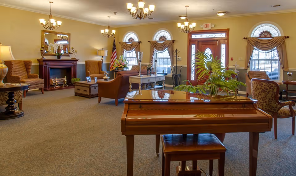 Spacious common room with a polished wooden piano in the foreground, upholstered chairs, a fireplace, chandeliers, and the front entrance.