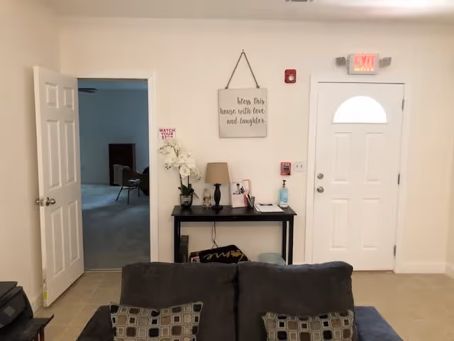 Interior view of a living room and entryway with a sofa in the foreground, a console table with decor beneath a wall sign, and a white front door.