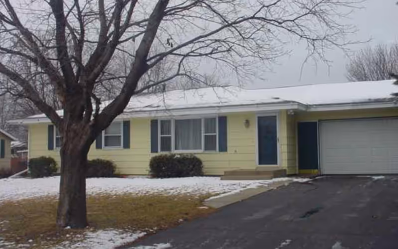 Single-story yellow facility exterior with blue shutters, garage, snow-covered lawn, leafless tree.