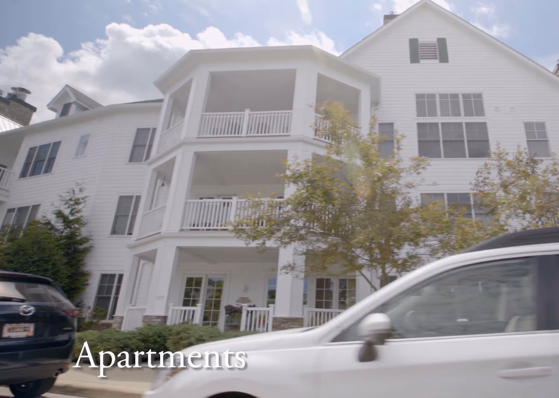 Front exterior of a white multi-story senior living building with stacked covered balconies and cars parked on the street.