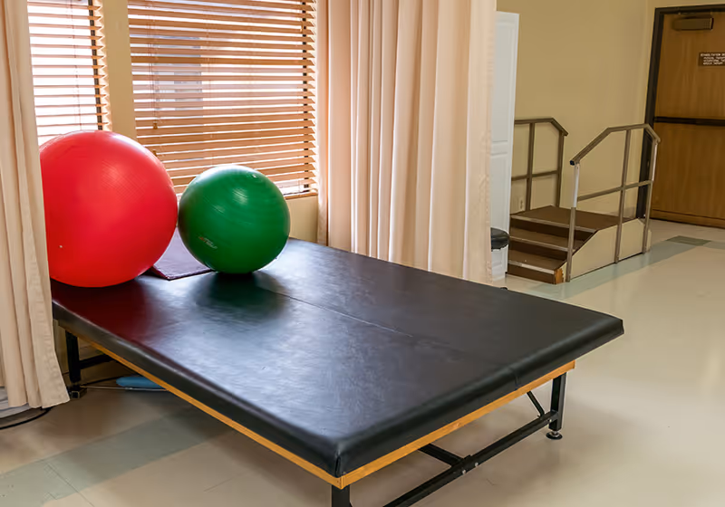 A therapy or examination room with a black padded treatment table, two exercise balls (one red and one green) on the table, beige curtains, a window with wooden blinds, and a small set of stairs with handrails leading to a door in the background.