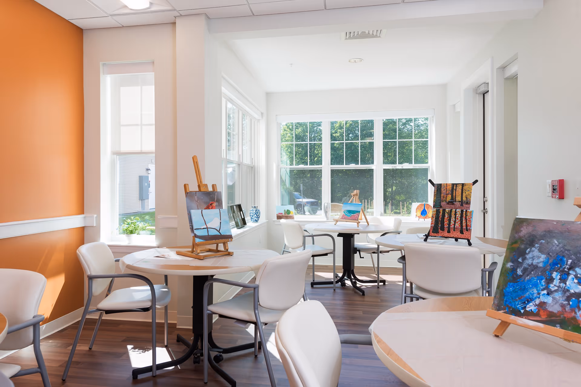 A bright and airy activity room with large windows letting in natural light. Several round tables with white chairs are arranged around the room, each table displaying small easels holding colorful paintings. The walls are painted white with one accent wall in orange, and the floor is a dark wood finish.