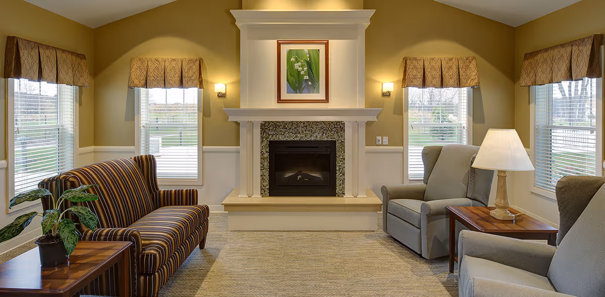 A cozy living room with a striped sofa on the left and two gray armchairs on the right, separated by wooden side tables with a potted plant and a lamp. The room features a central fireplace with a mosaic tile surround and a framed picture above it. Four windows with beige valances let in natural light, and the walls are painted a warm beige color.