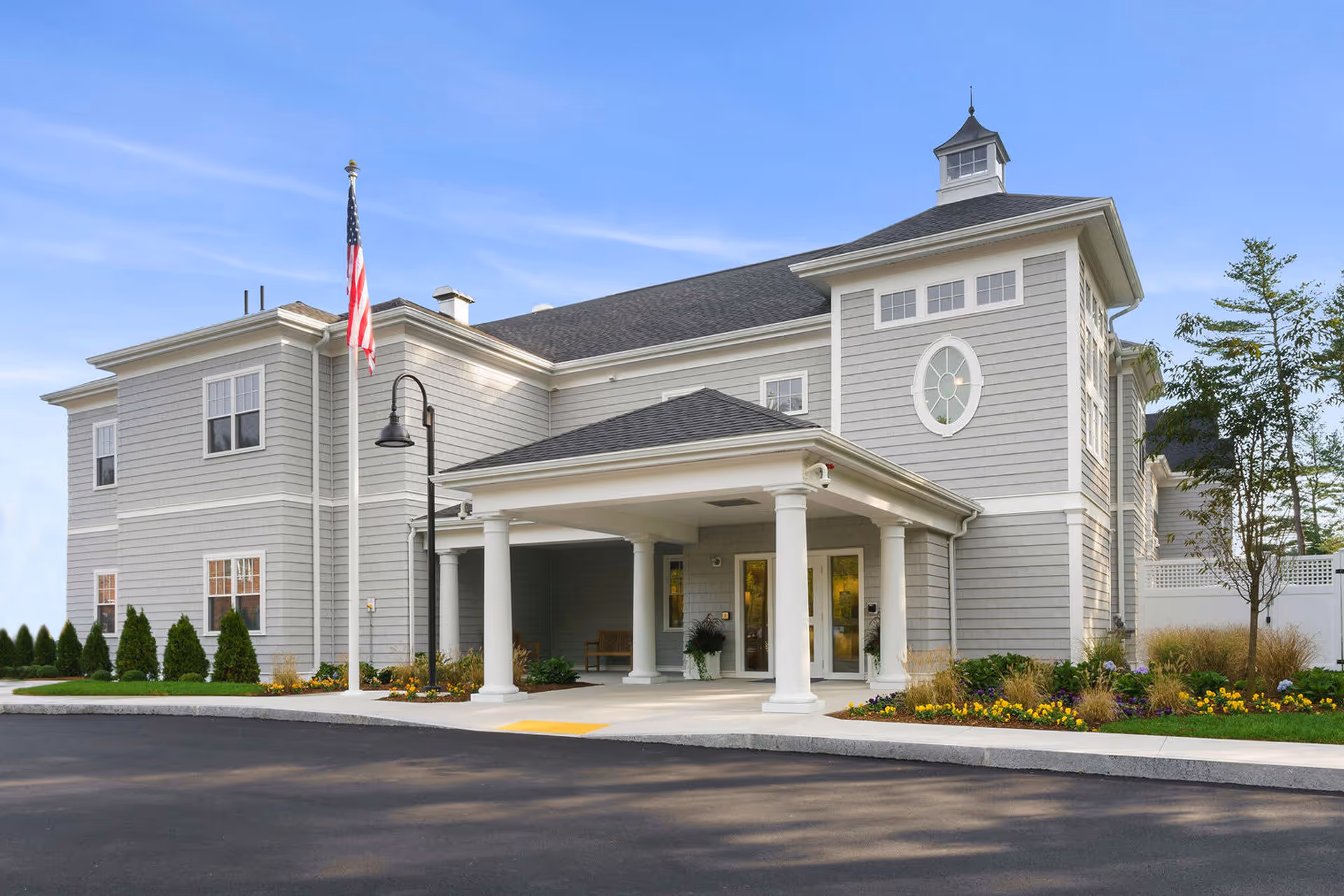 Exterior view of a two-story gray building with white trim, featuring a covered entrance supported by white columns, an American flag on a flagpole, landscaped flower beds, and a clear blue sky.