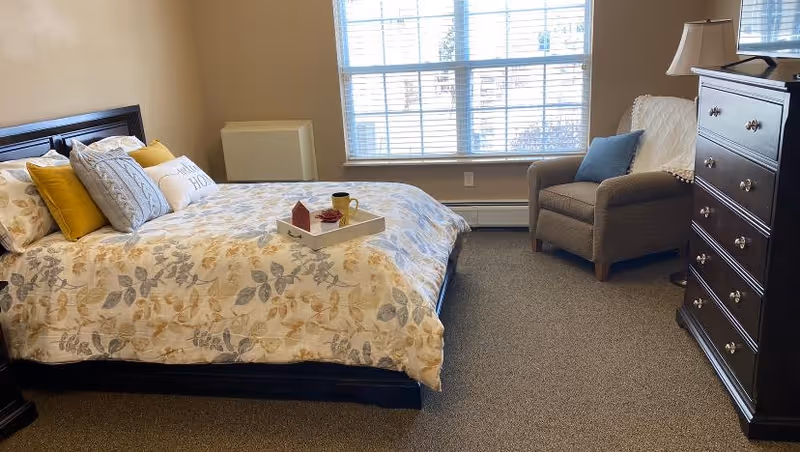 A cozy bedroom with a large bed covered in a floral patterned comforter and multiple pillows, including a decorative pillow that says 'This is Home'. A tray with a cup and small items is placed on the bed. Next to the bed is a cushioned armchair with a blue pillow and a white throw. A tall dark wooden dresser with a lamp on top stands against the wall. A large window with blinds allows natural light to fill the room.