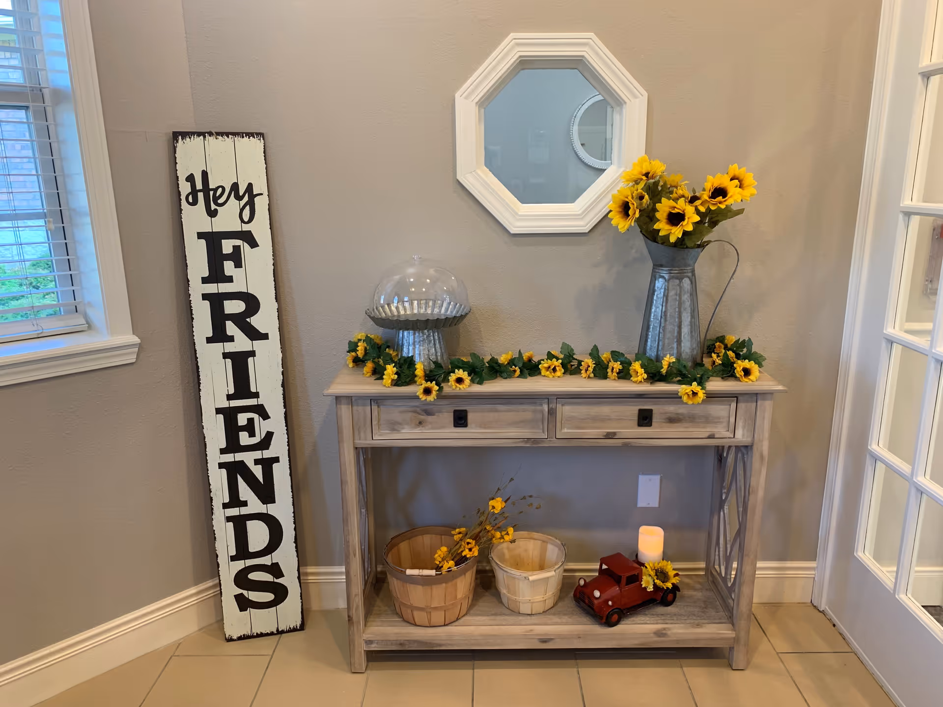 A decorative wooden console table against a beige wall with a small octagonal mirror above it. The table is adorned with a garland of yellow flowers, a metal pitcher filled with sunflowers, a glass cloche, two wooden baskets, a small red toy truck with a sunflower, and a battery-operated candle. To the left of the table is a tall wooden sign that reads 'Hey FRIENDS' in black letters. There is a window with blinds on the left and a glass-paneled door on the right.