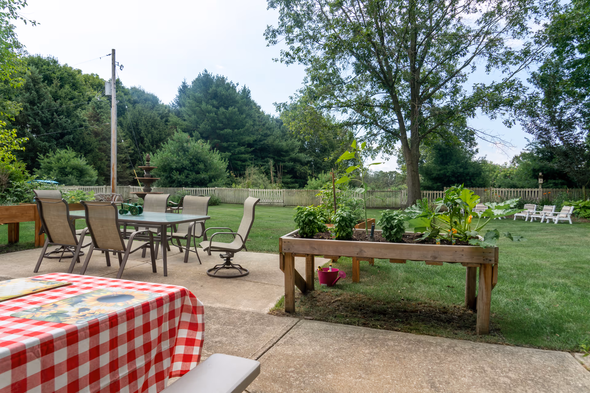 Patio with an outdoor dining table, raised garden beds, and lawn chairs overlooking a grassy yard and trees.